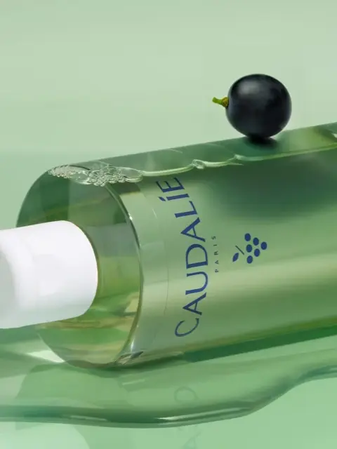 Green Caudalie bottle with white cap, grape and liquid on green surface.