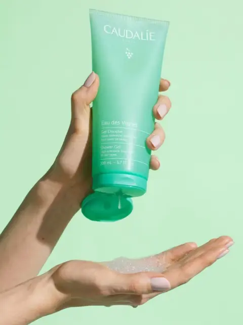 Hand holding Caudalie shower gel, dispensing onto palm against a light green background.