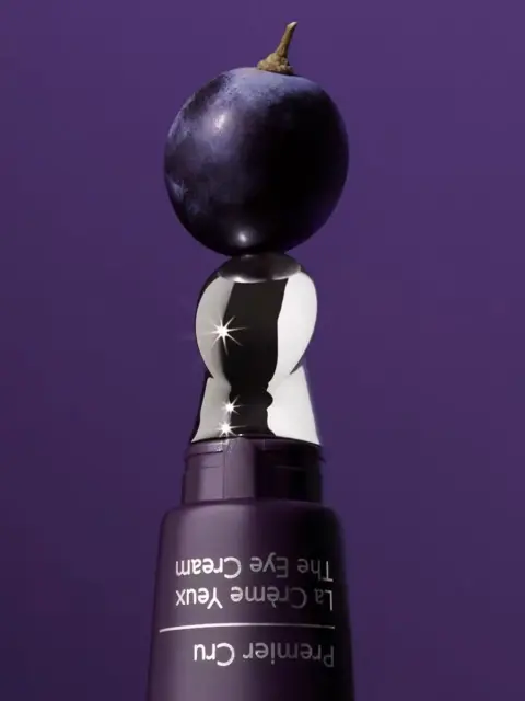 Purple eye cream bottle with a shiny silver cap, topped by a grape, on a purple background.