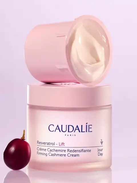 Caudalie Resveratrol Lift cream jar with lid off, grape beside, on a light purple background.