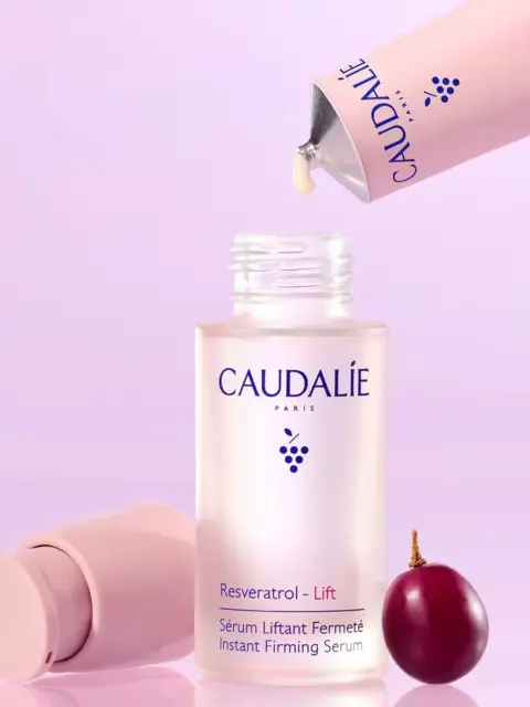 Caudalie Resveratrol-Lift serum bottle with grape and pink cap on purple background.