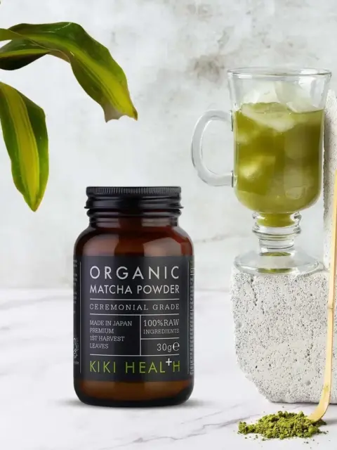Kiki Health organic matcha powder jar beside a glass of matcha drink and green leaves.
