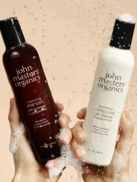 Two hands holding John Masters Organics shampoo and conditioner bottles with water droplets.