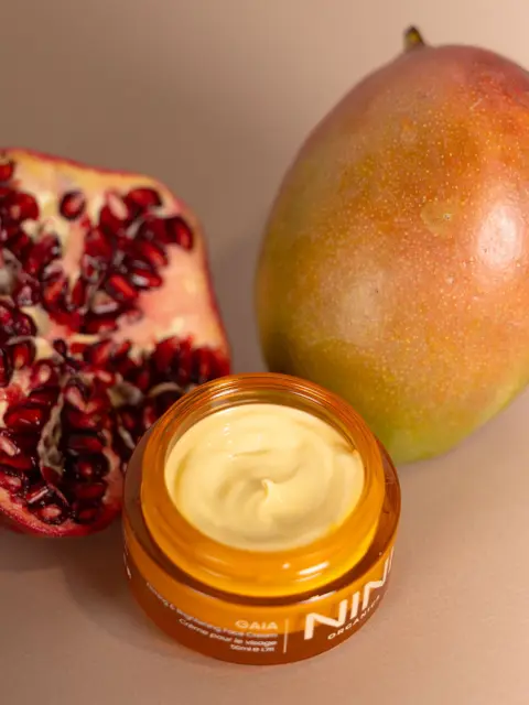 Open jar of cream with pomegranate half and whole mango on beige background.