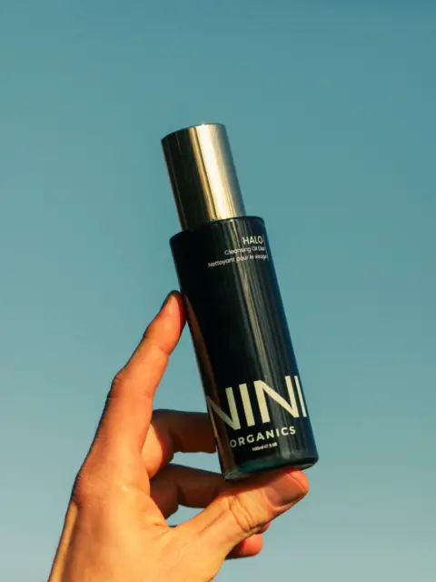 Hand holding NINI Organics bottle against a clear blue sky.