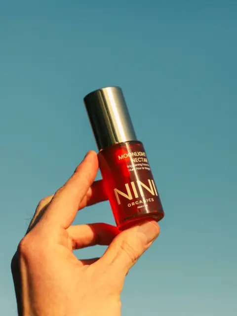 Hand holding NINI Organics Moonlight Nectar bottle against clear blue sky.