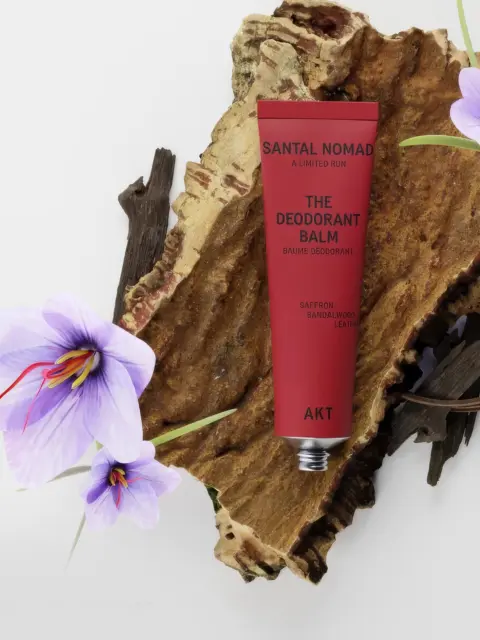 Red deodorant balm tube on bark with purple flowers. Text: Santal Nomad, AKT, saffron, sandalwood, leather.