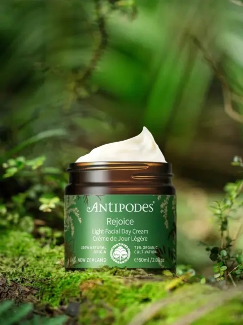 Antipodes Rejoice Light Facial Day Cream jar on a lush green forest floor.