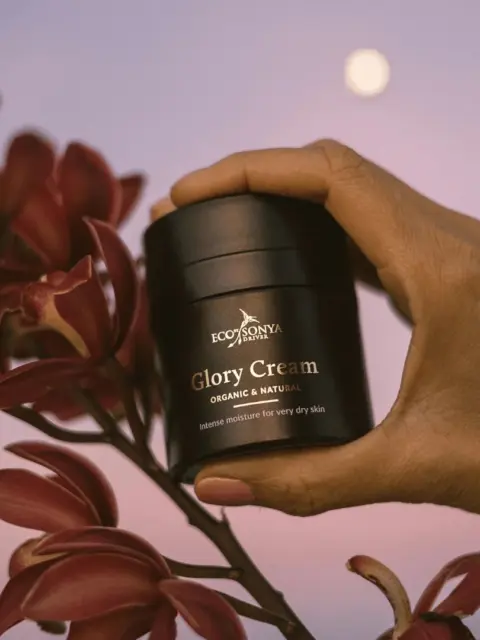 Hand holding Eco by Sonya Glory Cream near red flowers, moon in background.