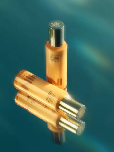Three orange skincare bottles with silver caps on a reflective blue surface.