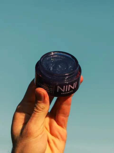Hand holding a NINI Organics jar against a blue background.