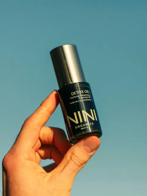 Hand holding NINI Organics Detox Oil bottle against clear blue sky.
