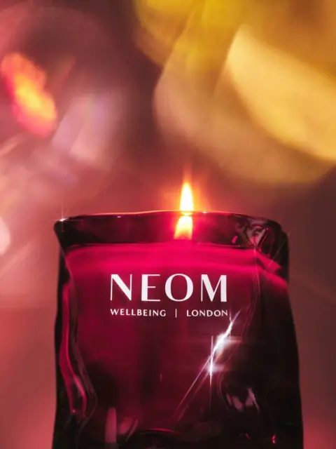 Red candle with flame, NEOM Wellbeing London text, blurred colourful background.