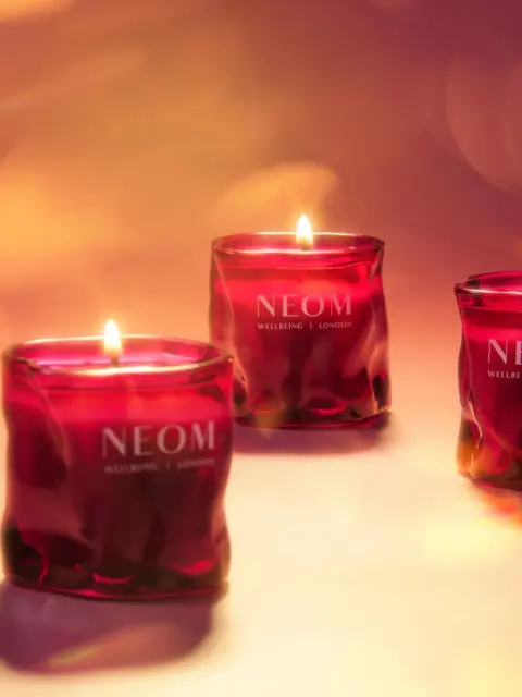 Three lit red candles with "NEOM Wellbeing London" text on a soft-focus background.