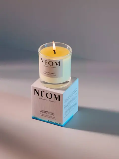 Lit NEOM candle on a box with "Scent to De-Stress" text, casting a shadow on a smooth surface.