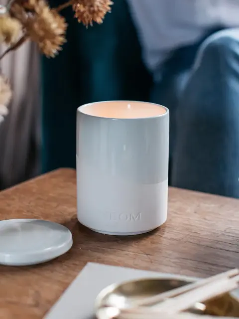 Lit white candle in a ceramic holder on a wooden table, with a lid beside it.