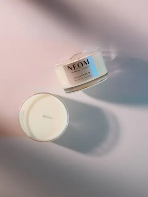 Two NEOM candles on a soft pink surface, one open showing the wick.