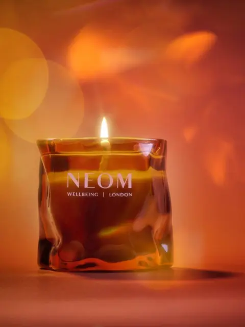 Lit NEOM candle with amber glass, text "Wellbeing | London", warm bokeh background.