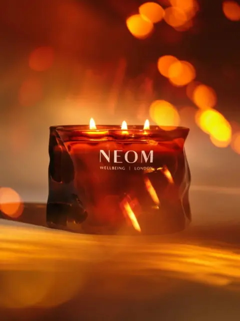 Lit candle in a dark amber glass with "NEOM Wellbeing London" text, warm glowing bokeh background.