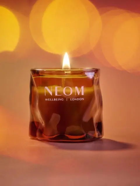Lit NEOM candle with "Wellbeing London" text, set against a warm, blurred background.