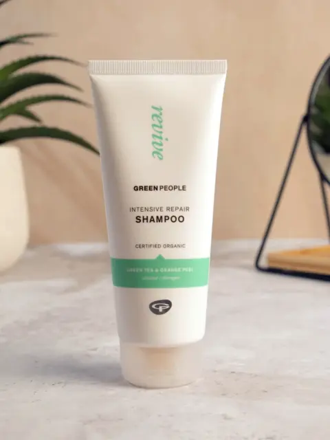 Green People Intensive Repair Shampoo tube on a bathroom counter with plants and a mirror.