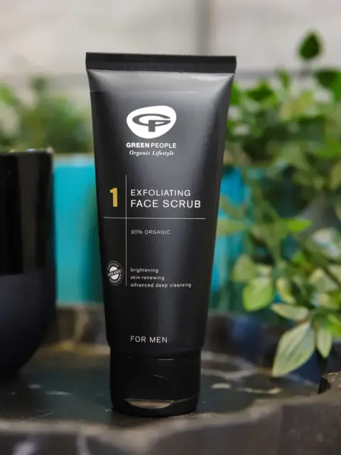 Black tube of Green People exfoliating face scrub for men, 90% organic, on a table with plants.