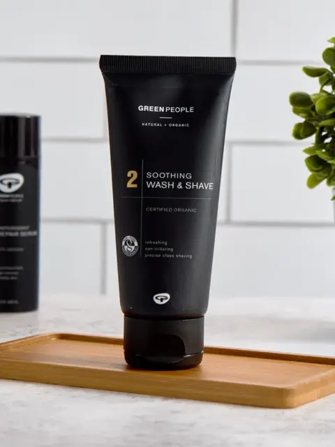 Black tube of Green People Soothing Wash & Shave on a wooden tray, plant in background.