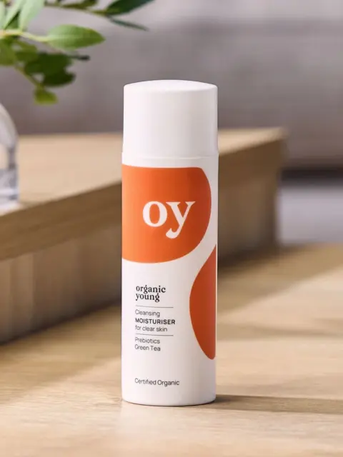 Organic Young moisturiser bottle on wooden surface, plant in background.