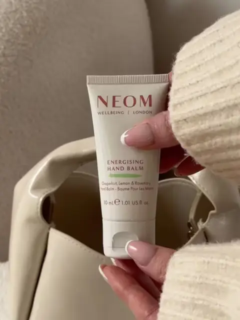 NEOM Energising Hand Balm, 30ml, held by a person in a cream jumper, near a beige bag.