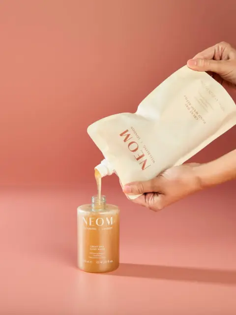 Hands pouring liquid from refill pouch into NEOM hand wash bottle.