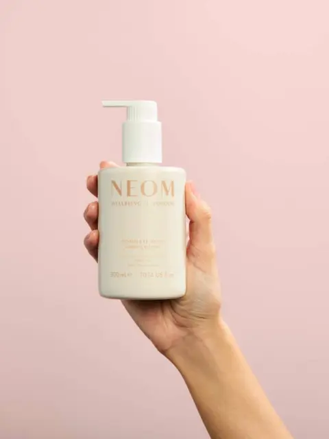 Hand holding NEOM lotion bottle against a pink background.