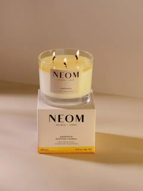 NEOM Happiness scented candle on box, lit with three wicks, in soft lighting.