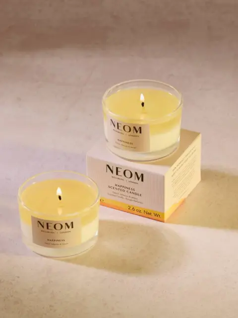 Two lit NEOM candles, one on a box, on a light surface.