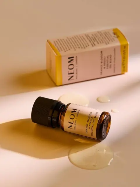 Neom essential oil bottle with box, oil spilled on a beige surface.