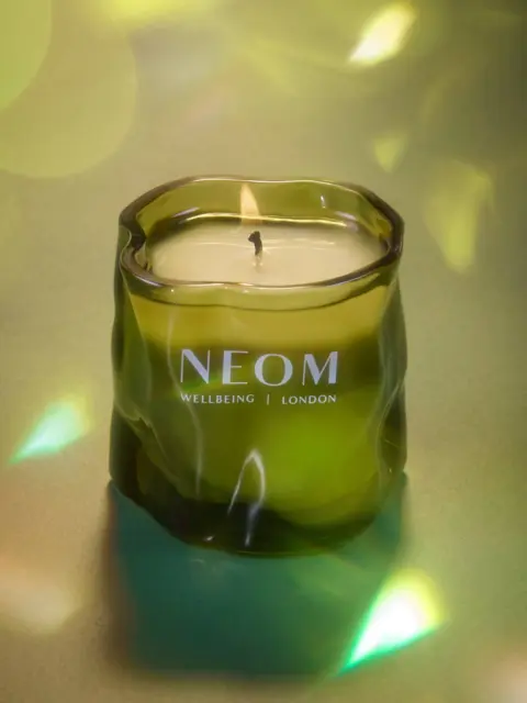 Green glass candle with "NEOM Wellbeing London" text, lit wick, on a reflective surface.