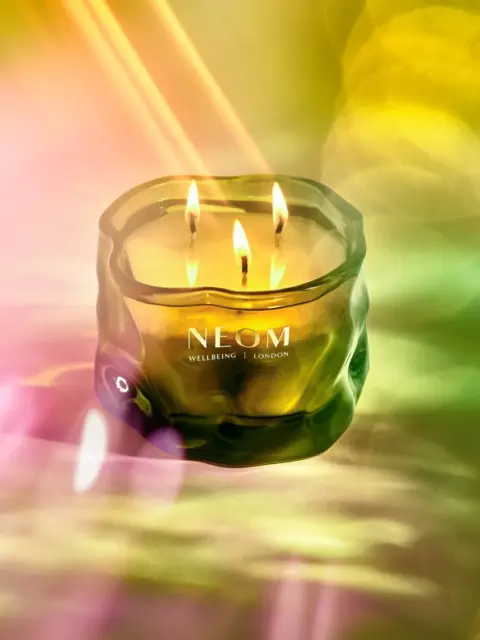 Green glass candle with three flames, labelled "NEOM Wellbeing London" against a colourful background.