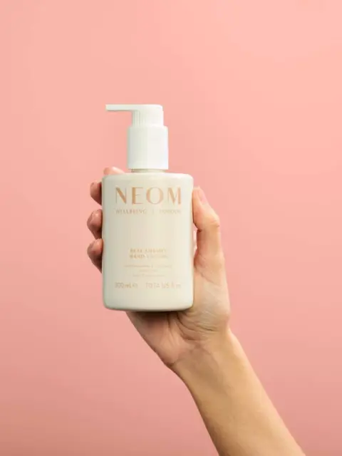 Hand holding NEOM hand lotion bottle against pink background.