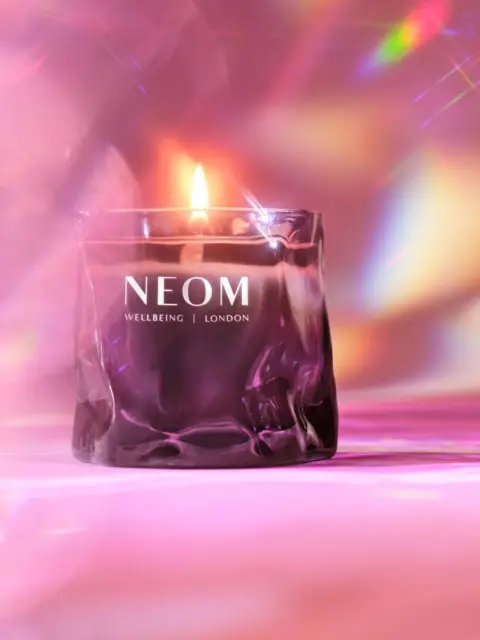 Lit candle in a glass holder with "NEOM Wellbeing London" text, pink and colourful background.
