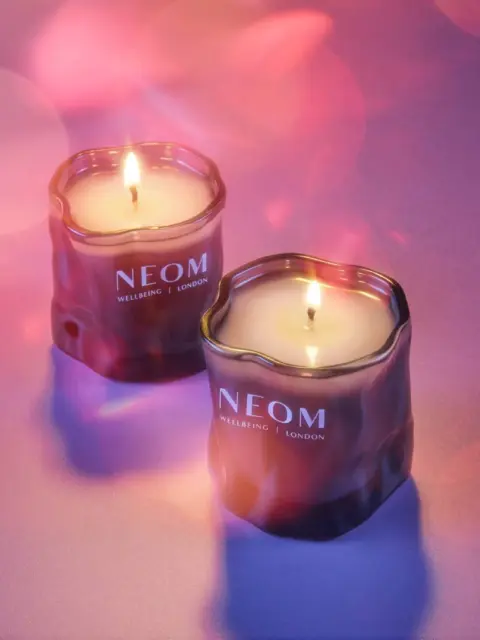 Two lit NEOM candles on a purple surface with soft pink bokeh lights.