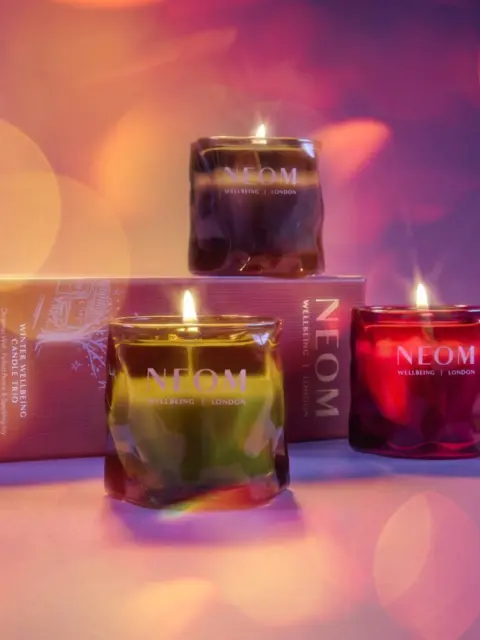 Three lit NEOM candles in green, brown, and red glass jars with a branded box behind.