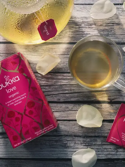 Pukka Love organic tea with teapot, cup, rose petals, and card on wooden surface.