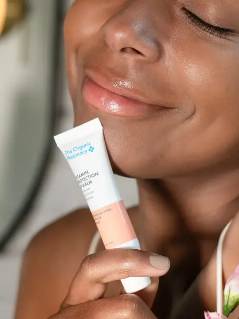 Woman holding The Organic Pharmacy lip balm, smiling with closed eyes.