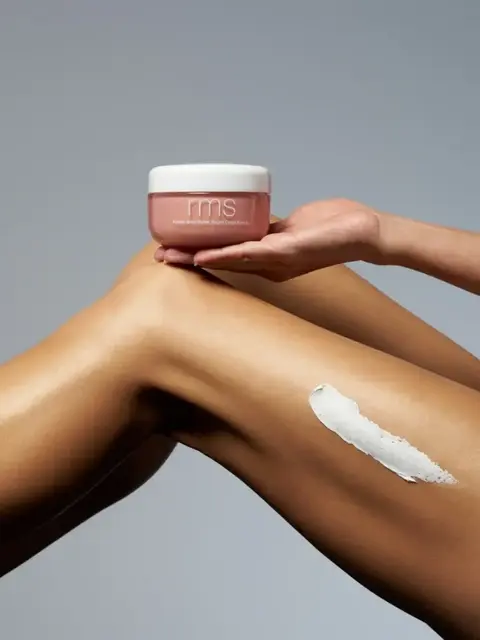 Hand holding RMS body butter above a leg with cream applied.