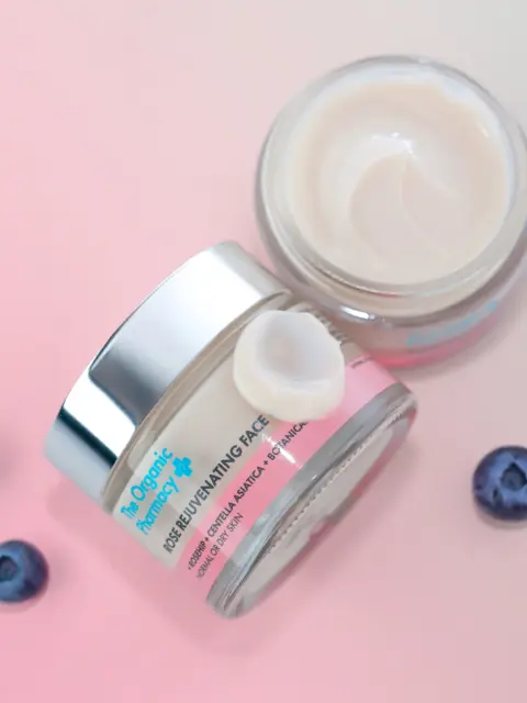 Two jars of rose rejuvenating face cream on a pink surface with blueberries.