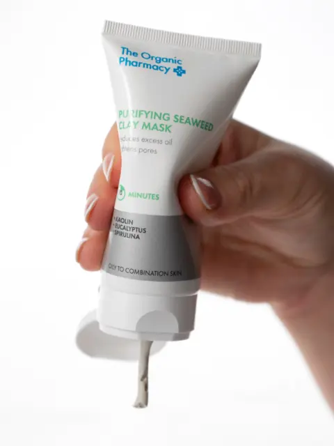 Hand holding The Organic Pharmacy clay mask tube, dispensing product.