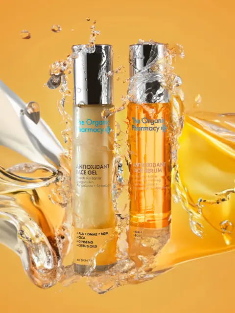 Two bottles of The Organic Pharmacy antioxidant gel and serum with liquid splash on orange background.
