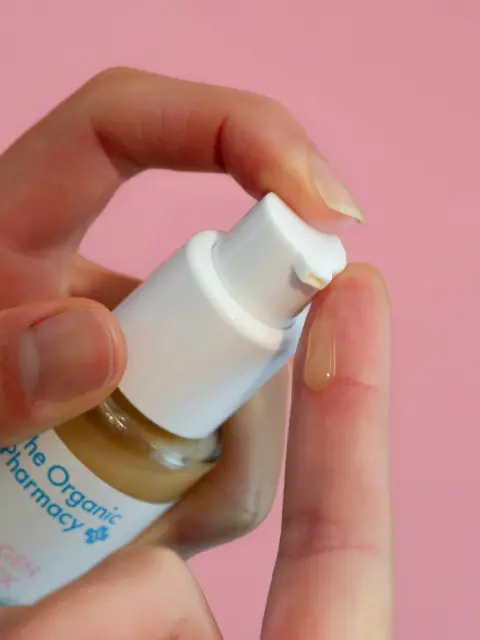 Hand dispensing serum from a white pump bottle onto a fingertip. Pink background.