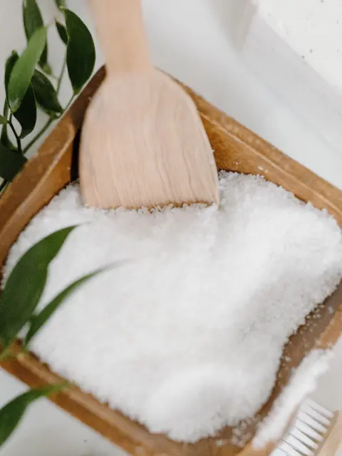 Wooden bowl of bath salts with a scoop, green leaves, and a bar of soap nearby.