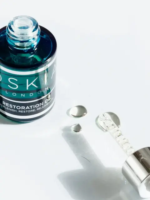 Open bottle of Oskia Restoration Oil with dropper and oil droplets on white surface.
