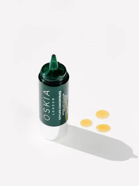 Oskia London skincare bottle with three yellow serum drops on white background.